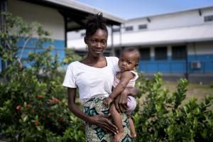 woman stands outside holds her daughter in arm and looks toward the camera