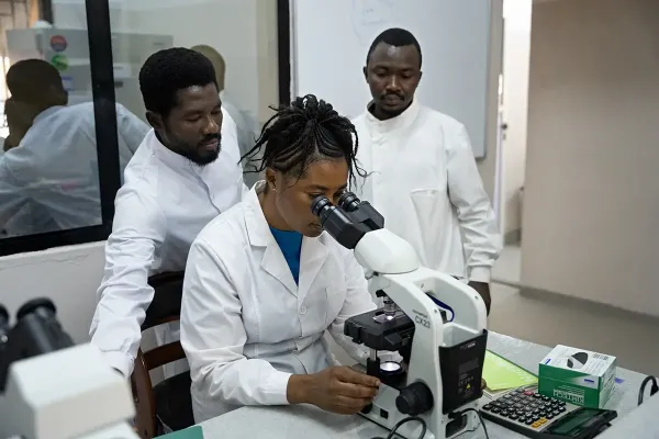 woman is looking through microscope in lab, two men standing next to her
