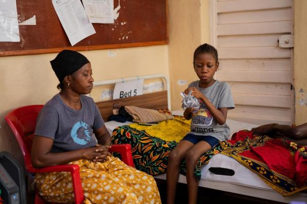Mariatu drinks water from a plastic bag while sitting on the hospital bed, her mother sitting by her side.