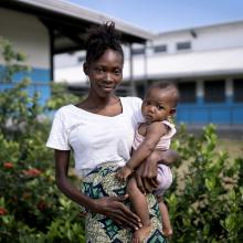woman stands outside holds her daughter in arm and looks toward the camera