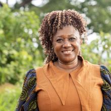 A smiling woman with braided hair stands outdoors against a background of green trees and foliage.