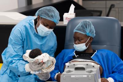 nurse holding newborn wearing personal protective equipment sitting next to woman in NICU.