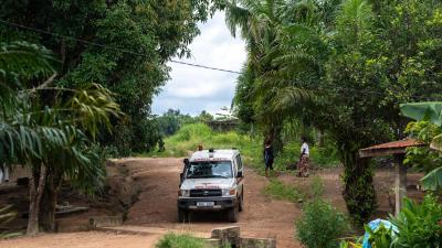 An ambulance navigates a dirt road surrounded by green trees