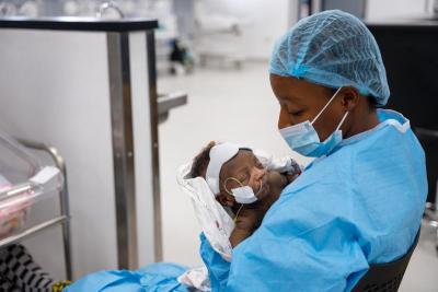 A sitting woman in a blue medical gown, cap, and face-mask holding a newborn baby with a breathing tube.