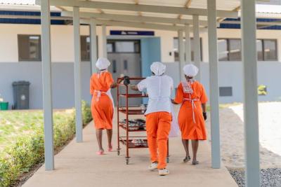 Hind view of two women and and a man in orange and white attire pushing a four-tiered trolley down a covered walkway.