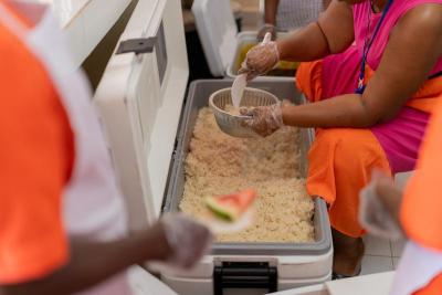 A sitting woman's gloved hands adding rice to a metal bowl from a large tub of rice.